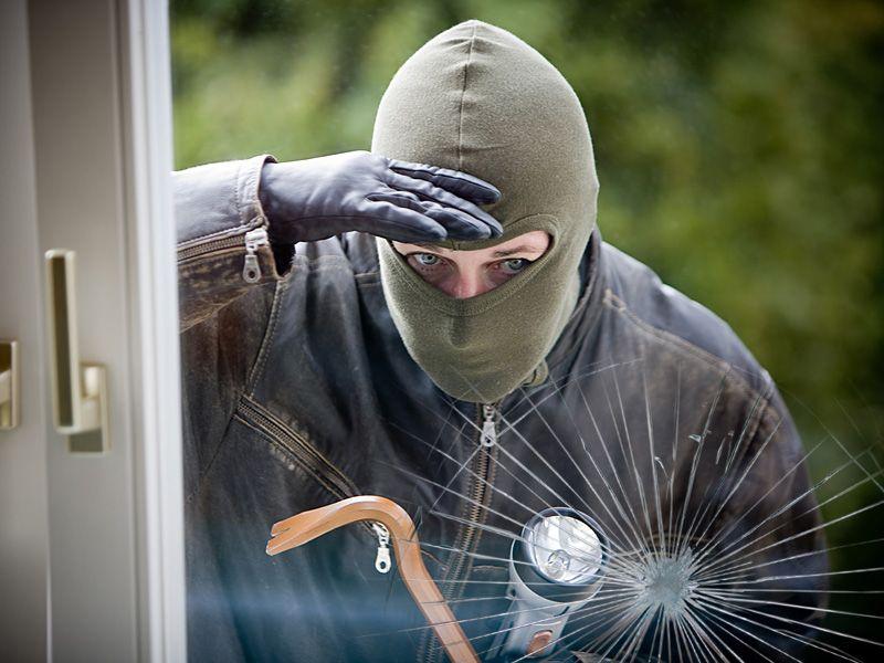 Ein Einbrecher mit Maske und Handschuhen schaut durch ein zerbrochenes Fenster